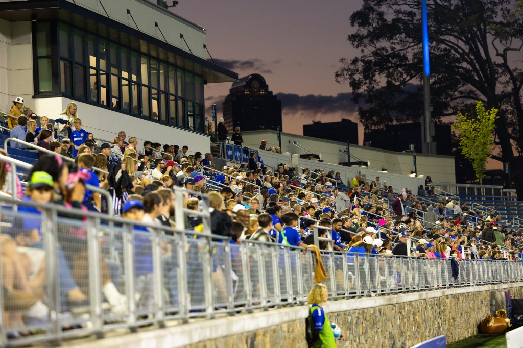 View of the crowd at a Charlotte Independence game at American Legion Memorial Stadium. The stands and press box are visible at an angle with the stone wall at the base. The sun has set and the fans are illuminated by bright stadium lights.