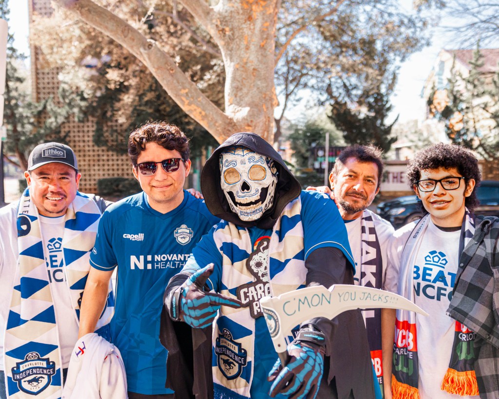 A group of five men standing on the sidewalk with a tree behind them. Three of the men are wearing white t-shirts that say "Beat NCFC" on them. The other two men are wearing blue Charlotte Independence jerseys. The man in the center is dressed in a reaper costume with a white skeleton mask and holding a scythe that says "C'mon You Jacks".