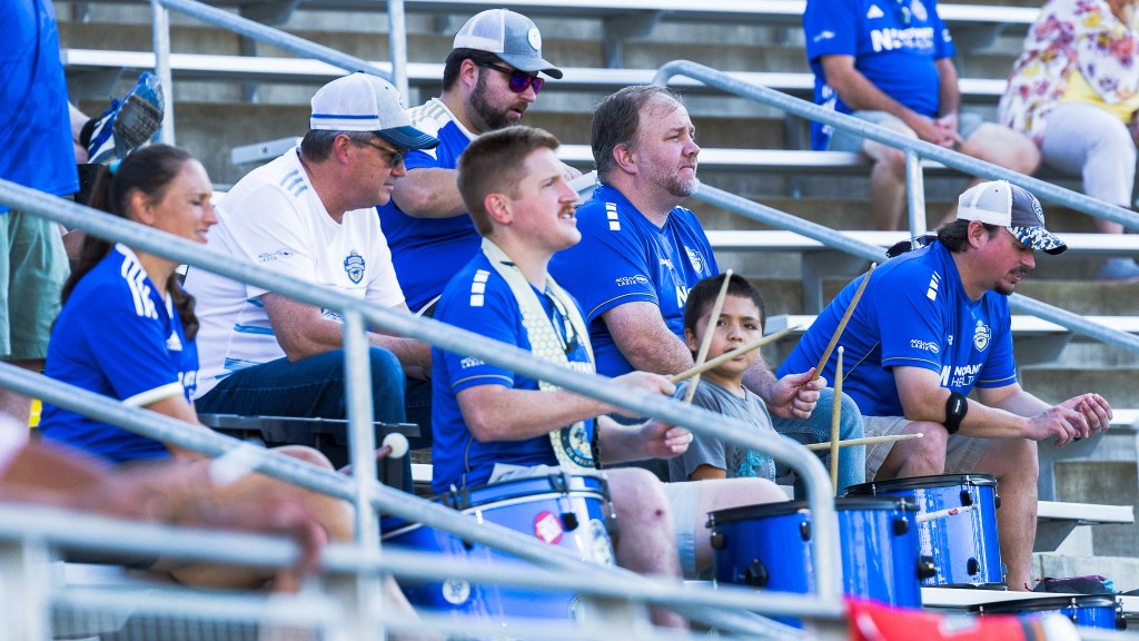Charlotte Independence fans dressed in blue jerseys drumming while sitting in metal bleachers.