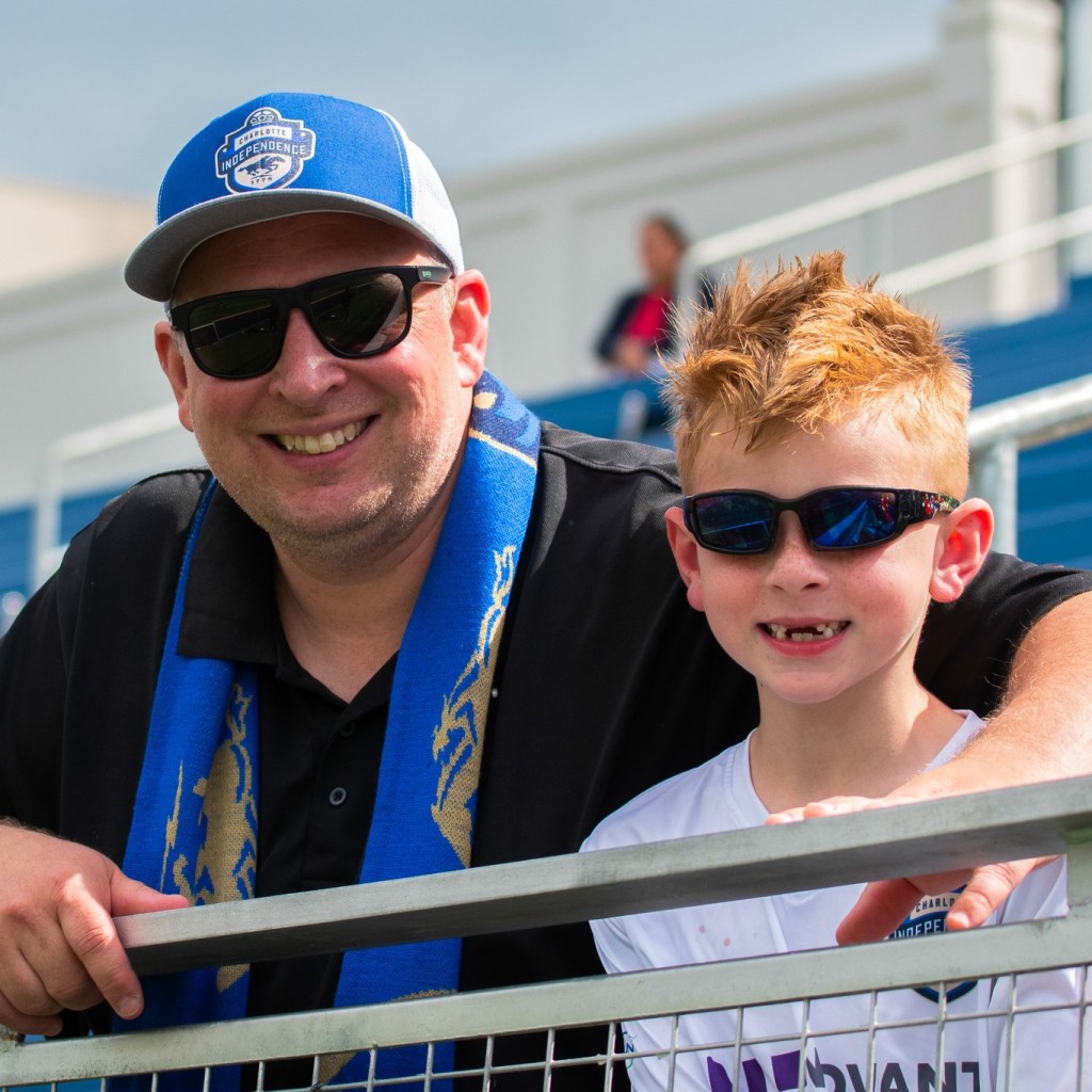 A dad and son smiling for a picture at a Charlotte Independence game. The dad is wearing a blue hat, sunglasses, black shirt, and blue scarf. The son has red hair and is wearing sunglasses and a white shirt.