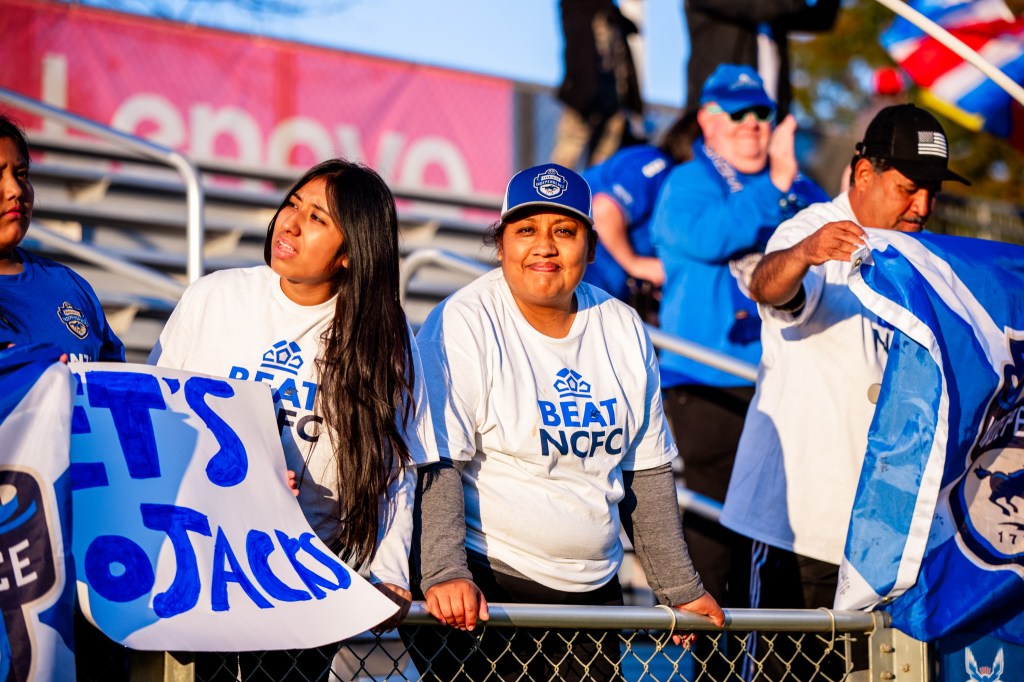 A picture of two women standing up against the railing of the stands at WakeMed Soccer Park. Both women are wearing white shirts with text that says "Beat NCFC". The woman on the left has dark brown hair and is holding a white sign with blue letters that say "Let's Go Jacks". The woman on the right has dark brown hair and is holding the railing below her.