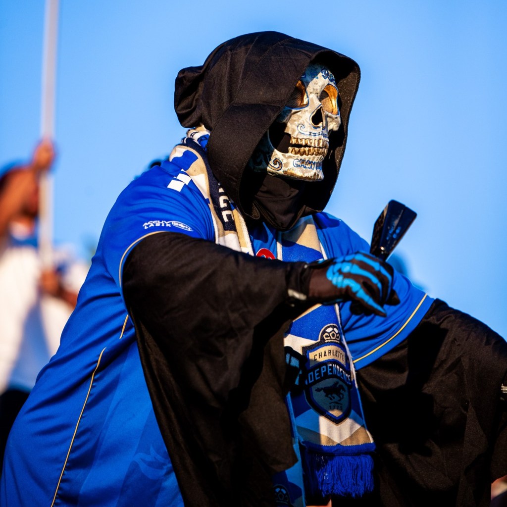 A man wearing a reaper costume with a blue Charlotte Independence jersey over top it leaning over slightly with a blue sky behind him. He is also wearing a white skeleton mask and holding a bell in his right hand.