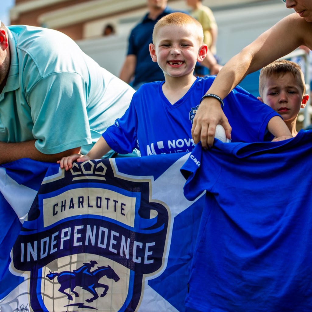 A small child with red hair wearing a blue shirt smiling while standing behind a railing with a Charlotte Independence flag hanging over it.