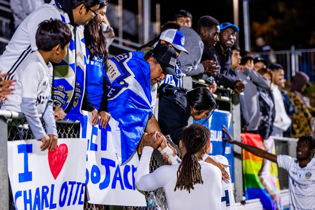 A view of the Charlotte Independence fans at WakeMed Soccer Park at the 2023 USL League One Final. Joel Johnson, a player for the Charlotte Independence is wearing a long-sleeve white undershirt while shaking the hand of a fan in the stands.
