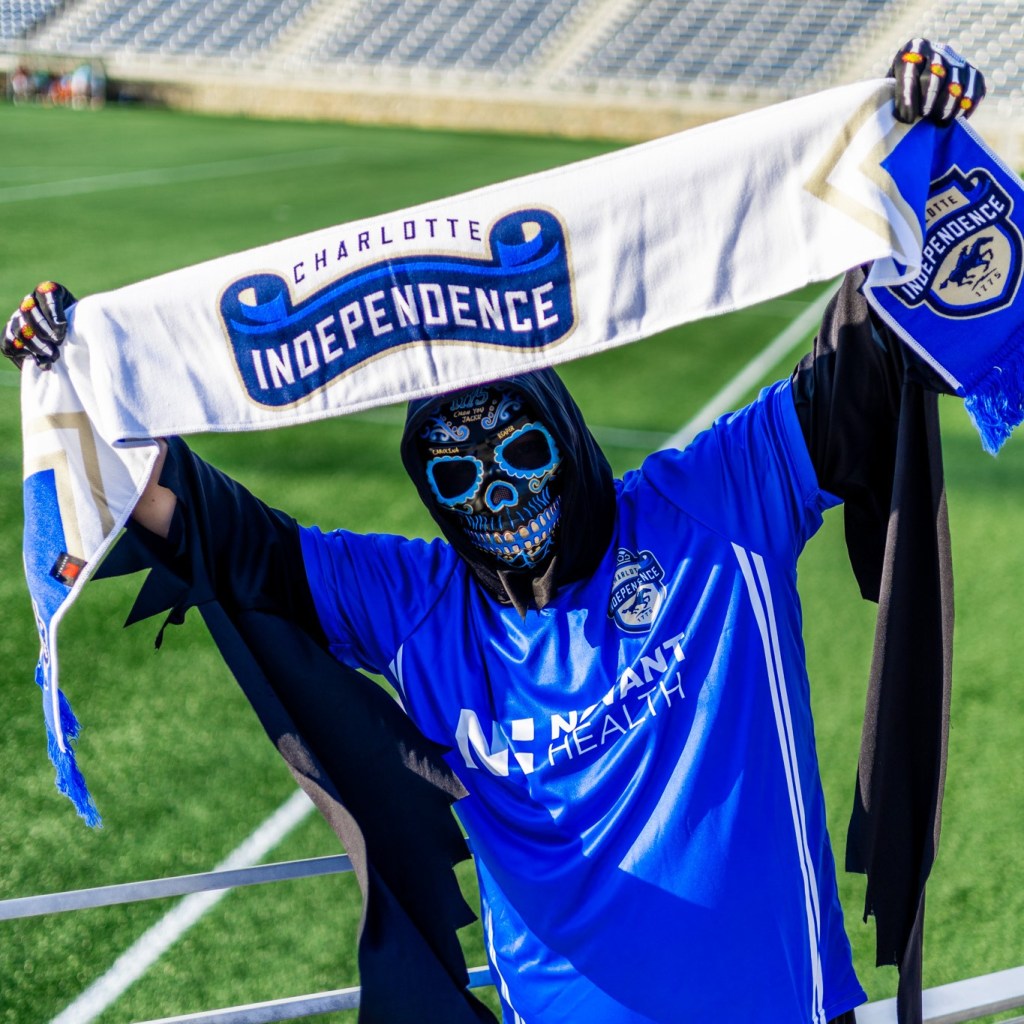 A man dressed in a reaper costume wearing a blue Charlotte Independence jersey over top it standing in front of the field at American Legion Memorial Stadium. He is also wearing a blue skeleton mask while holding an outstretched white Charlotte Independence scarf.