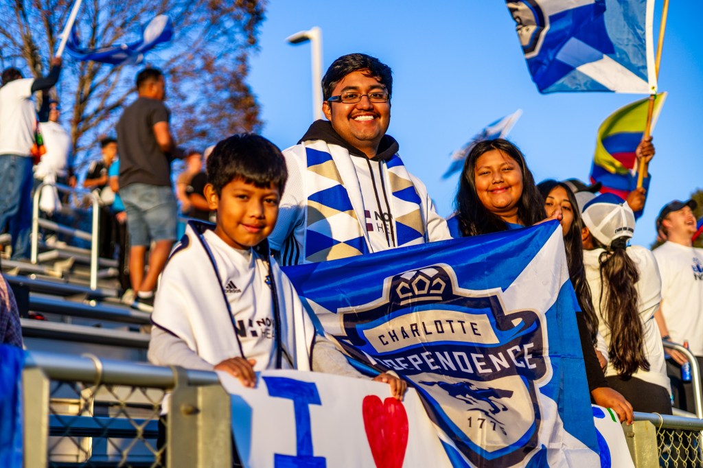 A group of three people standing in the bleachers at WakeMed Soccer Park. The boy on the left has dark brown hair and is wearing a white shirt while holding a white sign that says "I heart Charlotte". The man in the middle has dark brown hair and glasses. He is wearing a white shirt with a patterned gold, blue, and white scarf on top. He is holding a Charlotte Independence flag over the railing. The girl on the right is hidden mostly by the flag. She has dark brown hair.