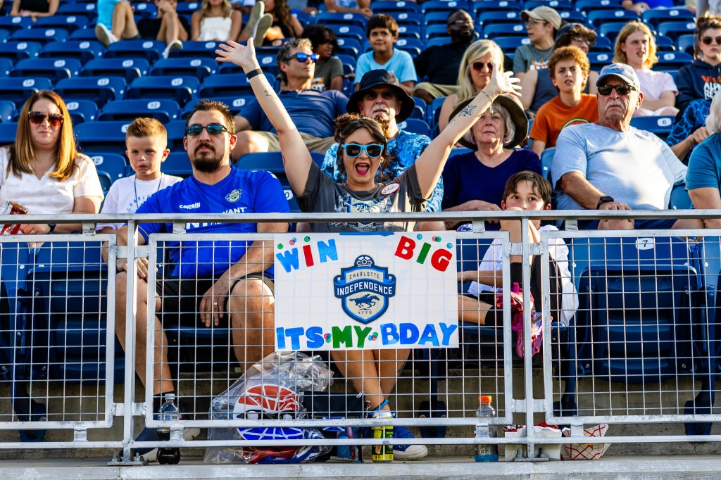 A picture of fans sitting in the blue chair back seats in the stands at the Charlotte Independence game at American Legion Memorial Stadium. The main focus of the picture is a woman sitting in the front row wearing blue sunglasses and a gray shirt with her hands in the air. On the railing in front of her, there is a sign that says "Win Big, It's My B-Day" with the Charlotte Independence logo in the center.