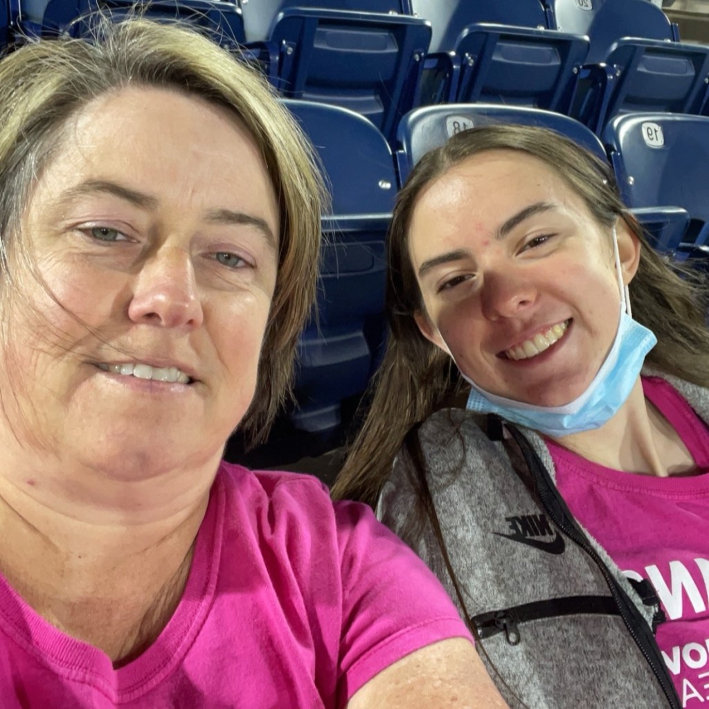 A selfie of two women wearing pink shirts sitting in the stands at a Charlotte Independence game. The woman on the right is wearing a gray jacket over the shirt and has a face mask under her chin.