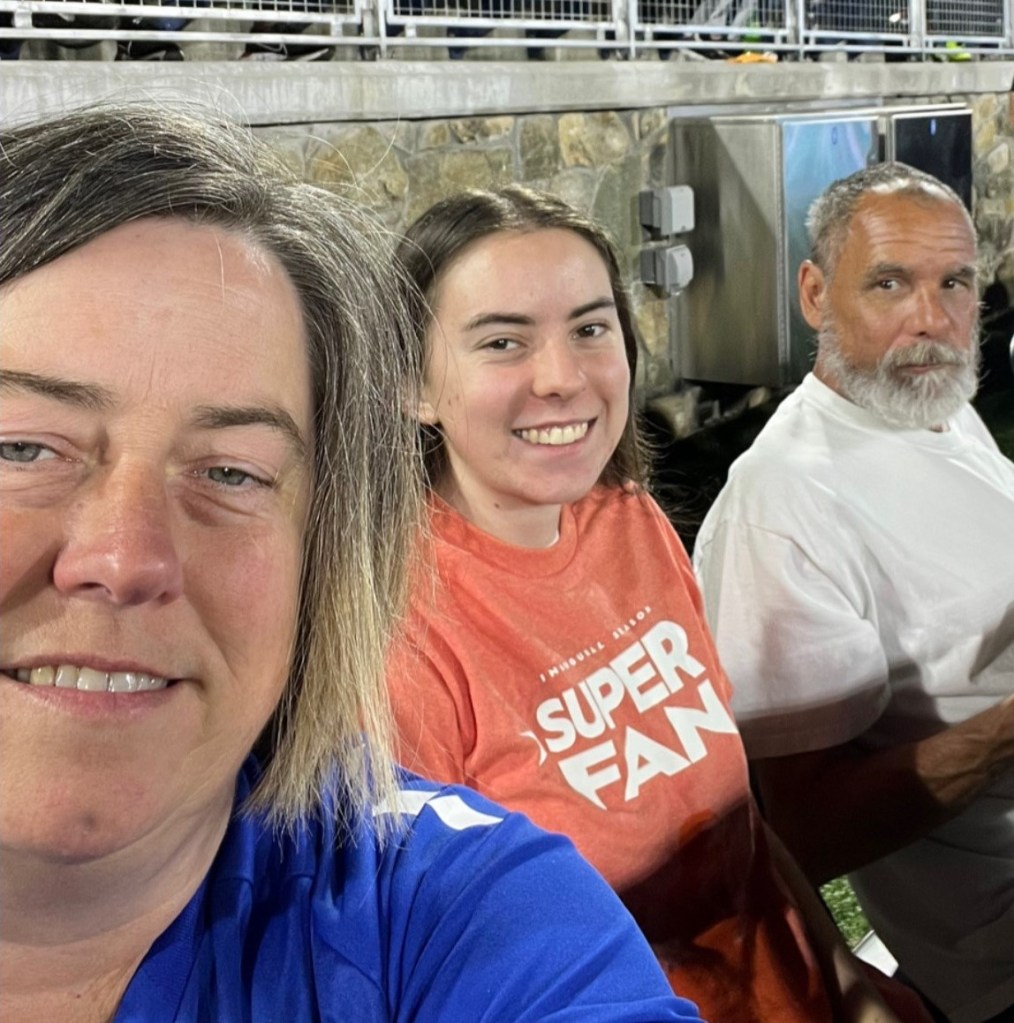 A selfie of three people sitting on the sideline at a Charlotte Independence game. The woman taking the picture is wearing a blue shirt next to a younger girl in an orange shirt that says "Super Fan". The third person is a gentleman wearing a white shirt.