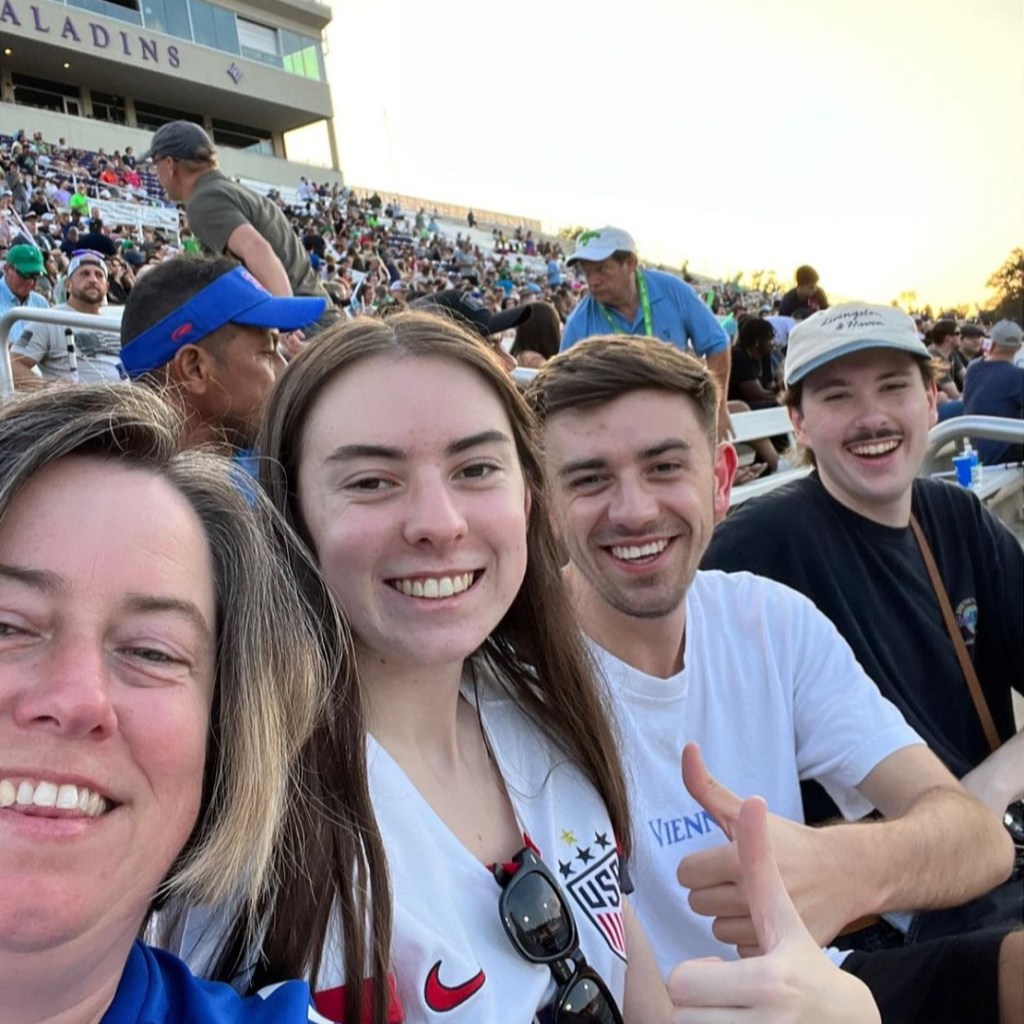 A selfie of four people at a Charlotte Independence game at Paladin Stadium. The woman taking the picture is wearing a blue shirt while sitting next to a younger girl wearing a white shirt with sunglasses hanging off the shirt. She is next to a guy in a white shirt giving a thumbs up. The gentleman sitting on the end is wearing a white hate and black shirt.