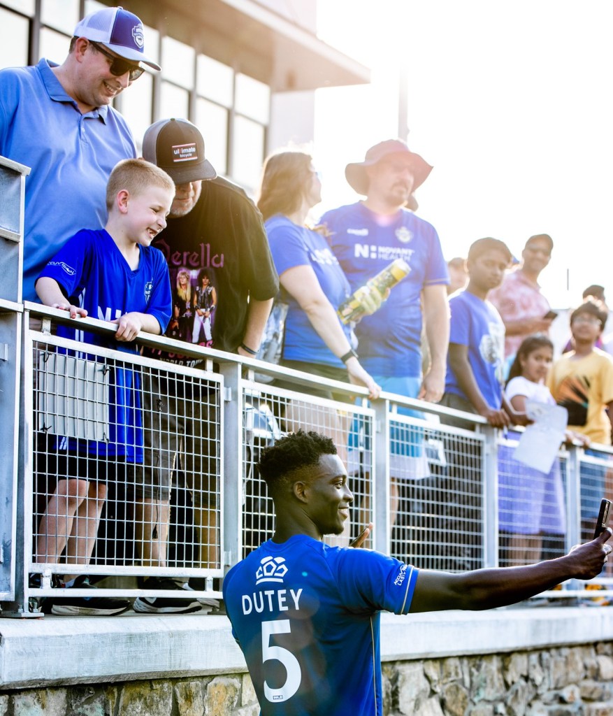 A crowd shot showing multiple people standing in the stands at a Charlotte Independence game. Charlotte Independence player #5 Shalom Dutey is standing below the stands taking a picture with a group of fans in the stands.