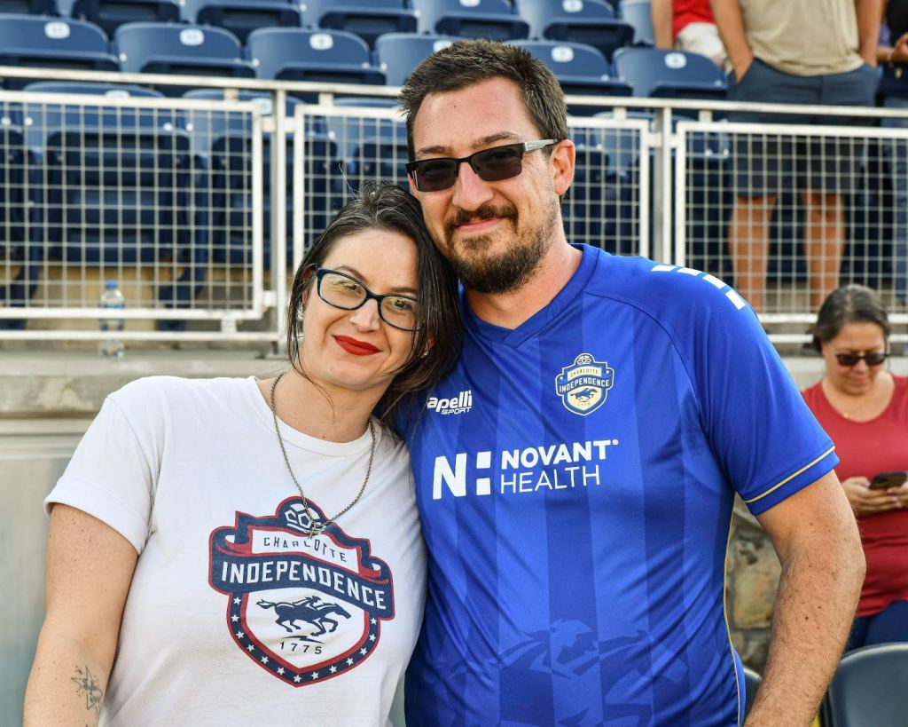 A woman standing next to a man on her left on the sideline of a Charlotte Independence game. She is wearing glasses and a white shirt with the red, white, and blue Charlotte Independence logo. The gentleman is wearing a blue Charlotte Independence jersey and sunglasses.