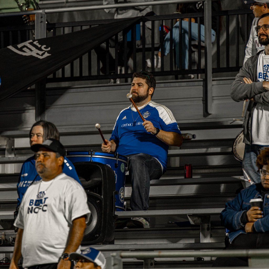 A man in a blue Charlotte Independence jersey playing a blue drum sitting in the bleachers at WakeMed Soccer Park at the 2023 USL League One Final. There are other people standing in the bleachers on the edges of the photo.
