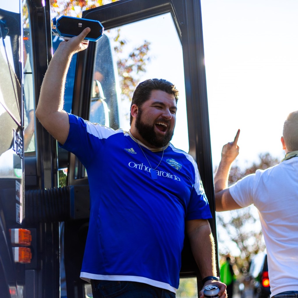 A man in a blue Charlotte Independence jersey with brown hair and a brown beard holding a portable speaker above his head in his right hand. He just stepped off a charter bus and is in front of the bus door.