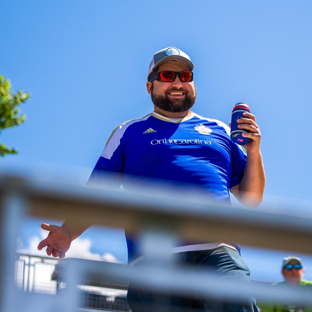 A picture of a man wearing a blue hat, red sunglasses, and a blue Charlotte Independence jersey. He is holding a beer can in his left hand. The railing of the bleachers is slightly obstructing the view of the man. He is standing in front of a blue sky.