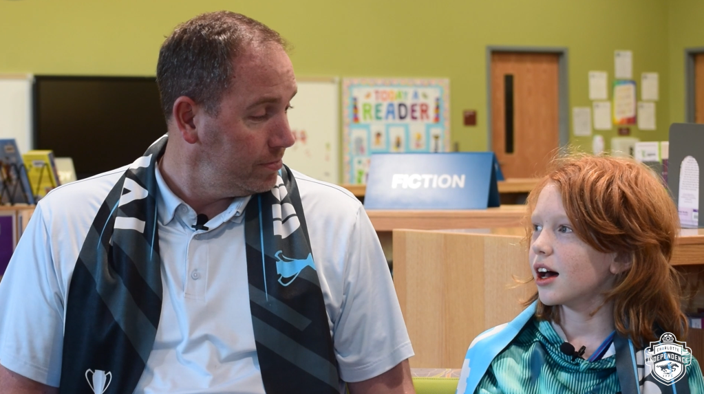 A father and son sitting in an elementary school library. Both of them are wearing a gray USL League One Fina; scarf. The dad on the left is wearing a pale blue polo. The son on the right is wearing a turquoise patterned hoodie.