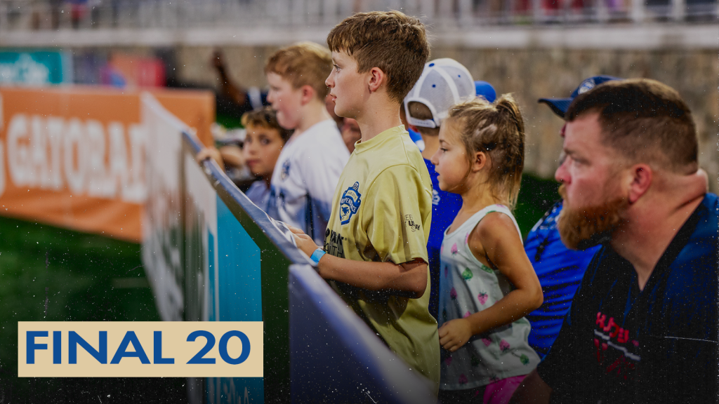 A photo of fans standing or sitting behind the field boards on the sideline at a Charlotte Independence game at American Legion Memorial Stadium. The subject of the photo is a young boy with blonde hair wearing a gold Charlotte Independence shirt standing against the field board. There is a gold text box in the bottom left corner with blue text that says "Final 20".
