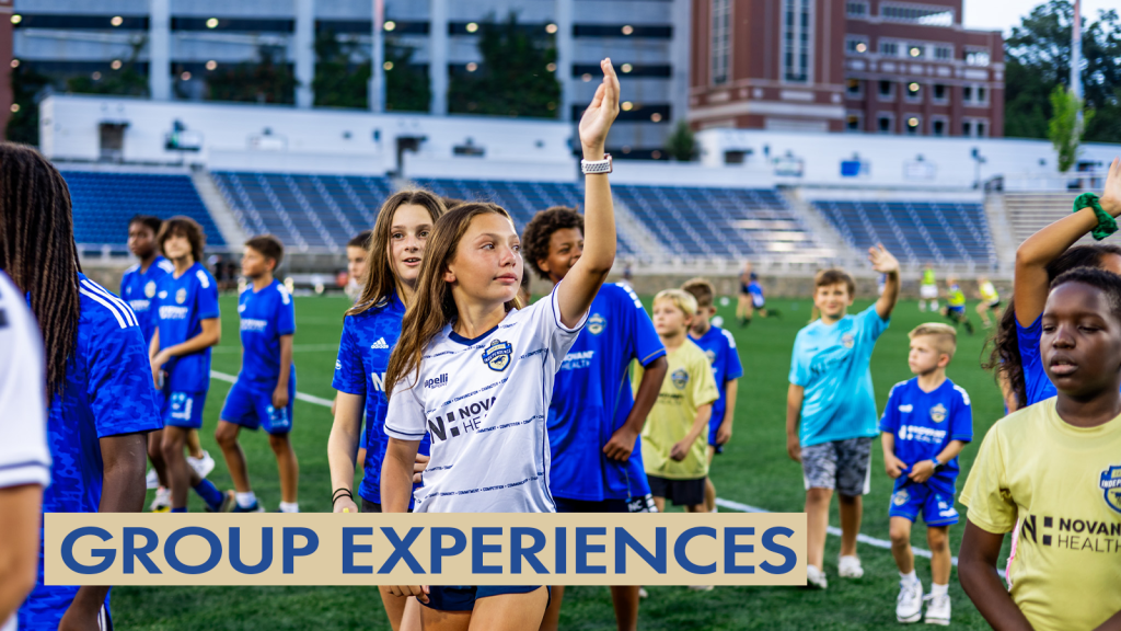Photo of a group of kids standing on the field at American Legion Memorial Stadium with the stands in the distance with the parking garage behind the stadium. All of the kids are wearing a Charlotte Independence shirt, either blue, gold, or white. The subject of the picture is a girl wearing a white jersey waving her left arm in the air while looking to her left. There is a gold text box in the bottom left corner with blue text that says "Group Experiences".