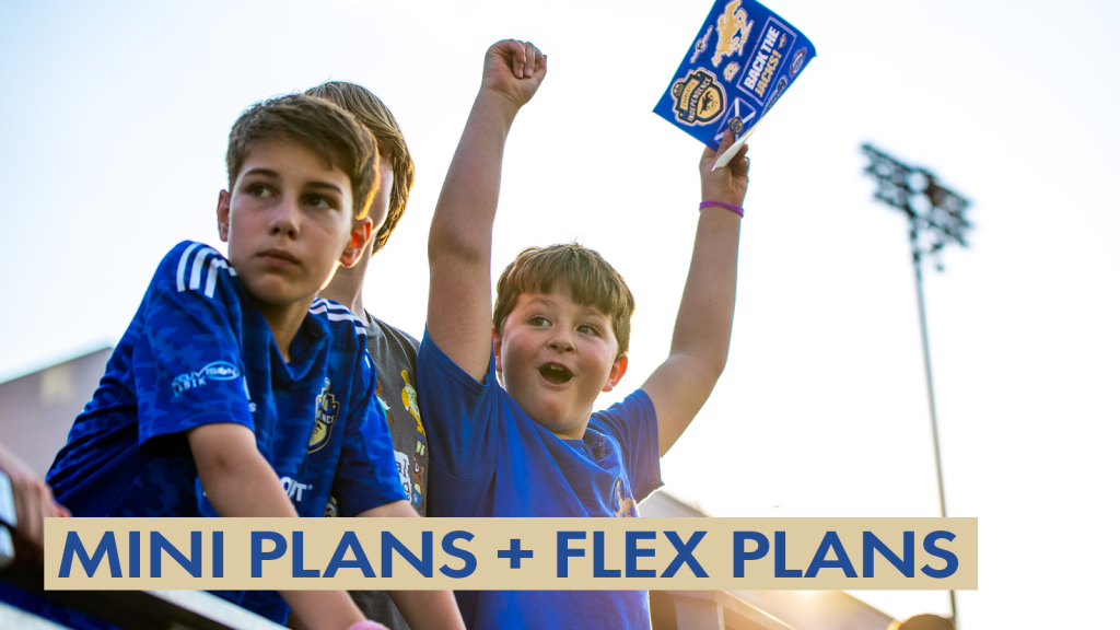 A phot of three boys in the stands at a Charlotte Independence game at American Legion Memorial Stadium. The subject of the photo is a young boy with brown hair wearing a blue shirt. He has both arms in the air and is holding a Charlotte Independence sticker sheet in his left hand. The boy in the middle is mostly hidden by the boy on the left who has brown hair and is wearing a blue jersey. There is a gold text box in the bottom left corner with blue text that says "Mini Plans + Flex Plans".