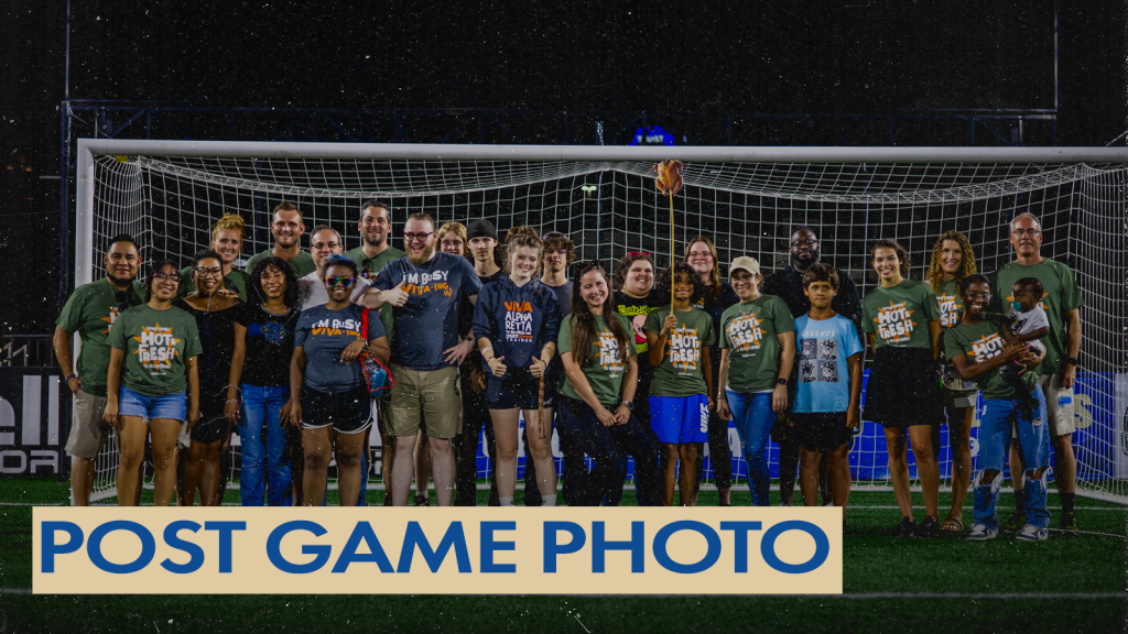 A group of adults standing in front of a soccer goal on the field at American Legion Memorial Stadium. Many of them are wearing an army green shirt that says Hot and Fresh with the Viva Chicken logo. There is a gold text box in the bottom left corner with blue text that says "Post Game Photo".