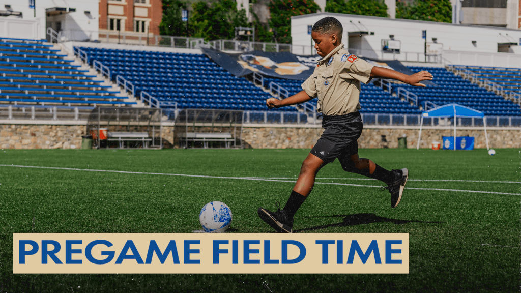 Photo of a young boy wearing a Scouts of American Uniform kicking a ball from the penalty spot at American Legion Memorial Stadium. The north seating is in the background. There is a gold text box in the bottom left corner with blue text that says "Pregame Field Time".