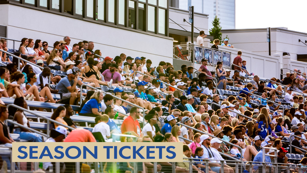 Photo of fans sitting in the stands at American Legion Memorial Stadium watching a Charlotte Independence game. There is a gold text box in the bottom left corner with blue text that says "Season Tickets".