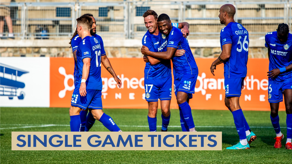 Photo of Charlotte Independence players wearing full blue uniforms standing on the field at American Legion Memorial Stadium in front of an OrangeTheory field board sign. #12 Brad Dunwell is walking in front of #8 Joel Johnson. To their right is #17 Clay Dimick hugging #90 Khori Bennett. #56 Gabriel Obertan is next to him, and #6 Omar Ciss is on the right edge of the frame. There is a gold text box in the bottom left corner with blue text that says "Single Game Tickets".